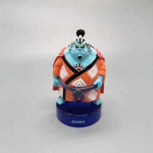 Puede incluir: Figura del personaje Jimbe de la serie de anime One Piece. La figura tiene piel azul, ropa naranja y blanca, y un peinado blanco y negro. Está sobre una base azul con el nombre "Jimbe" impreso.