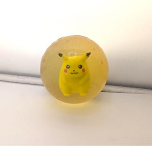 May include: A spherical, translucent, light yellow bouncy ball featuring a small, yellow Pikachu figure embedded in the center. The character has black-tipped ears, red cheeks, and a simple facial expression, encased within the clear material.