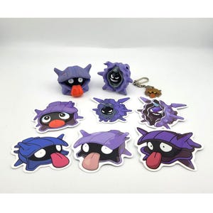 May include: A collection of purple and blue cartoon character figures and stickers. The figures have a shell-like appearance with black eyes and protruding tongues. A keychain with a similar character is also included.