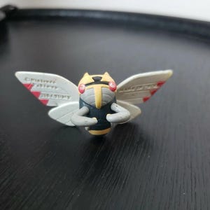 Pode incluir: Pequena e detalhada figura de um inseto estilizado com asas brancas, corpo amarelo e cinza e detalhes vermelhos. As asas têm texto e marcas vermelhas. A figura está sobre uma superfície escura.