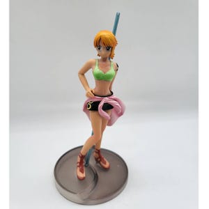 May include: A standing anime-style figurine of a character with short orange hair, wearing a light green sports bra, a black mini skirt with a tied pink garment around the waist, and brown sandals, positioned on a circular base with a blue pole.