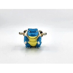 May include: A blue, yellow, and white toy figure of a turtle-like creature with two cannons on its back. The figure has a stern expression and is set against a white background.