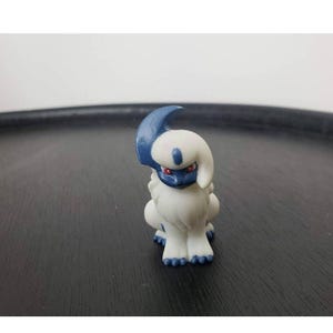 May include: A small, white and blue figurine of a creature with a curved horn, red eyes, and blue markings. The figurine is sitting on a dark surface.