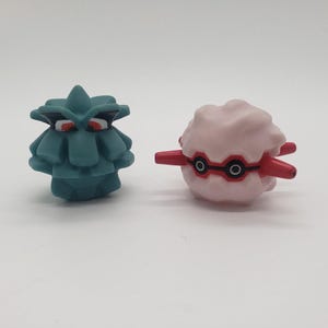 Peut inclure: Deux petites figurines en plastique de personnages Pokémon. La première est un Cloyster bleu et vert avec des yeux rouges. La seconde est un Forretress rose et rouge avec des yeux noirs.