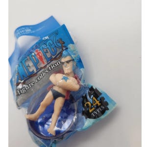 Puede incluir: Figura de acción de plástico de la colección "One Piece", empaquetada en una bolsa de plástico azul. La figura es de un personaje que lleva gafas de sol, una chaqueta roja y un bañador azul. El embalaje incluye el texto "24 TYPES".