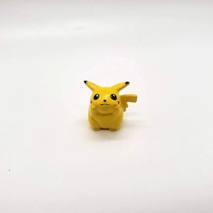 May include: A small, yellow Pikachu figurine with black-tipped ears, red cheeks, and a small tail. The figurine is a stylized representation of the popular Pokémon character, with a smiling expression. The figure is set against a white background.