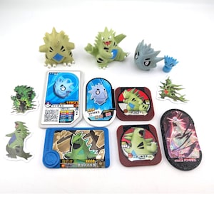 May include: A collection of Pokémon trading cards and figurines. The figurines are green and tan, depicting various Pokémon characters. The cards feature colorful illustrations of Pokémon, with Japanese text and QR codes. The items are arranged on a white surface.