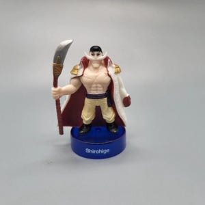 Puede incluir: Una figura de Barba Blanca de la serie de anime One Piece. La figura es musculosa, con un bigote blanco y un abrigo blanco con detalles en rojo y dorado. Sostiene una espada y se encuentra sobre una base azul con el nombre "Shirohige".