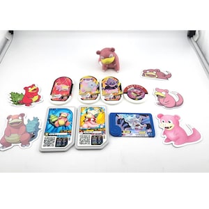 Op de afbeelding: Een verzameling Pokémon-ruilkaarten en beeldjes. De set bevat verschillende kaarten met kleurrijke illustraties van Pokémon-personages, samen met een klein, roze Slowpoke-beeldje. De kaarten bevatten Japanse tekst en QR-codes.