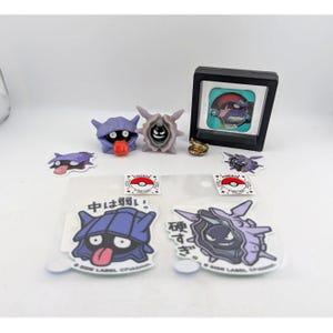 Op de afbeelding: Een verzameling Pokémon-items, waaronder stickers, beeldjes en een ingelijste afbeelding. De stickers tonen stripfiguren met Japanse tekst. De beeldjes zijn in verschillende kleuren en er is ook een kleine goudkleurige ring aanwezig.