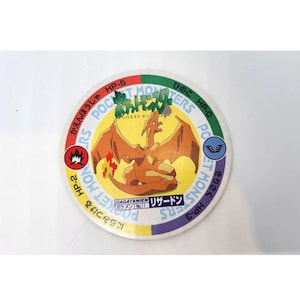 May include: Circular Pokémon trading card featuring Charizard, a fiery orange dragon. The card has Japanese text and a red, yellow, and blue border. The card also has the text "POCKET MONSTERS" and "HP-5".