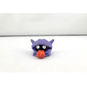 May include: A small, purple, cartoon-like figure with a black body, white eyes, and an orange tongue sticking out. The figure has a shell-like head and is set against a white background.