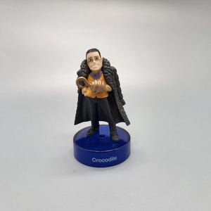 Puede incluir: Una figura de Crocodile de la serie de anime One Piece. La figura está de pie sobre una base azul con el nombre "Crocodile" impreso. Crocodile lleva un abrigo negro y sostiene un gancho.