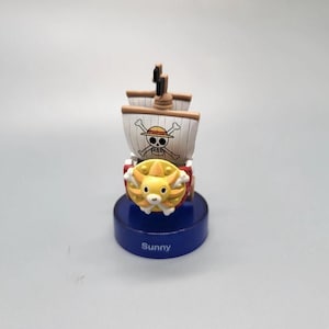 Puede incluir: Un pequeño barco de juguete con una figura amarilla con forma de león en la parte delantera y un mástil con un emblema de calavera y huesos cruzados. Las velas son beige y blancas. La base azul tiene la palabra "Sunny".