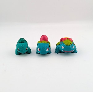 Puede incluir: Tres figuras de criaturas de color verde azulado dispuestas en fila. La primera es una criatura pequeña con un bulbo verde en la espalda. La segunda es una criatura con un capullo rosa. La tercera es una criatura más grande con una flor.