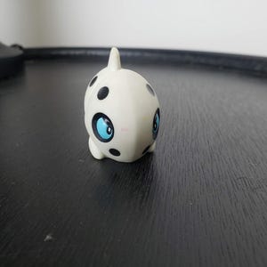 May include: A small, off-white figurine shaped like a stylized creature with a smooth, rounded body. It features prominent black polka dots scattered across its surface and large, expressive blue eyes with black pupils on either side of its head.