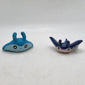 May include: Two small, blue manta ray figurines. One is light blue with a smiling face and the other is dark blue with a white belly. Both have curved horns and are likely handmade.