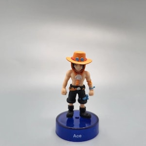 Puede incluir: Una pequeña figura del personaje Ace de la serie de anime One Piece. Ace lleva un sombrero beige con detalles naranjas, pantalones cortos negros y botas. La figura está montada sobre una base azul con el nombre "Ace".