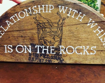May include: A wooden sign with the text "My relationship with whiskey is on the rocks". The sign is shaped like a half-circle and has a drawing of a glass of whiskey with ice.