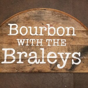 Puede incluir: Un letrero de madera con el texto "Bourbon with the Braleys" pintado en blanco sobre un fondo marrón.