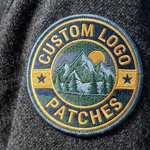 May include: A circular embroidered patch attached to a dark gray wool-like fabric. The patch features a mountain landscape with a yellow sun, green trees, and blue sky. The text CUSTOM LOGO PATCHES is stitched in yellow along the border.
