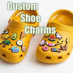 May include: Two yellow clogs decorated with colorful shoe charms. The charms include a rainbow, avocado, pizza slice, daisy, and a speech bubble that says "Have Fun". The text "Custom Shoe Charms" is at the top.