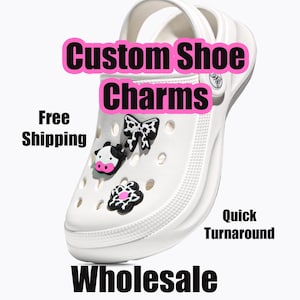 Può includere: Una scarpa a zoccolo bianca decorata con tre ciondoli bianchi e neri a forma di muso di mucca, un fiocco e un fiore. L'immagine include il testo Custom Shoe Charms, Free Shipping, Quick Turnaround e Wholesale. La scarpa ha un cinturino sul tallone.