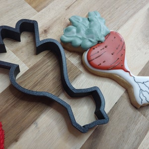 May include: A black plastic cookie cutter in the shape of a beet next to a decorated sugar cookie. The cookie is iced with red, white, and green frosting, and has a detailed design. The cookie cutter is on a wooden surface.