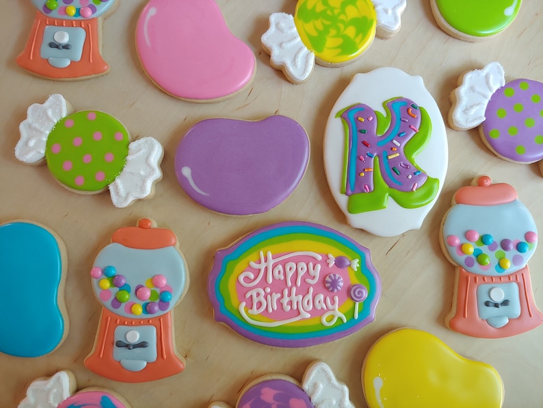 May include: Assortment of decorated sugar cookies in various shapes, including candy, gumball machines, and a cookie with the letter "K". The cookies are decorated with colorful icing and sprinkles, perfect for a birthday celebration.