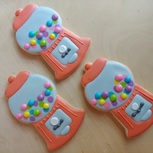 May include: Three gumball machine-shaped cookies. Each cookie is decorated with light blue icing for the main body, coral-colored accents, and colorful candy-shaped sprinkles. The cookies are arranged on a light wooden surface.