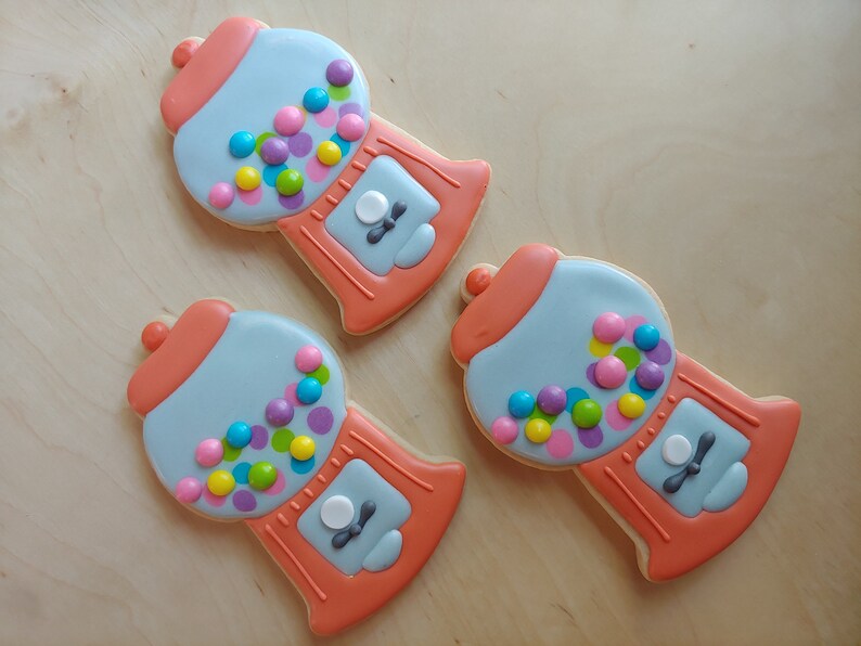 May include: Three gumball machine-shaped cookies. Each cookie is decorated with light blue icing for the main body, coral-colored accents, and colorful candy-shaped sprinkles. The cookies are arranged on a light wooden surface.