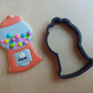 May include: A decorated gumball machine cookie with colorful candies and a matching cookie cutter. The cookie features orange, light blue, and pink icing with a variety of colorful candies. The cookie cutter is black.