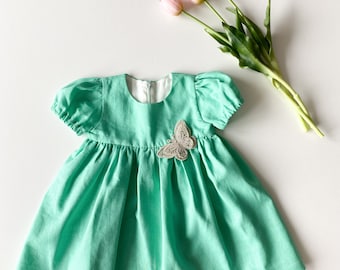 May include: A mint green linen dress with a butterfly applique. The dress has puffed sleeves and a gathered skirt.