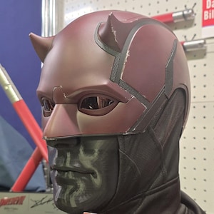 May include: A red and black Daredevil mask with horns and a black mask covering the lower face. The mask is made of plastic and has a textured surface.