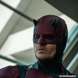 May include: A close-up of a person wearing a red and black Daredevil costume. The costume features a red mask with horns and a black suit. The person is looking to the right of the frame.