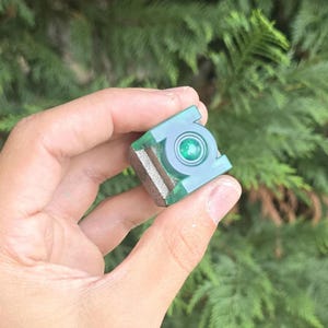 May include: A small, cube-shaped object featuring a translucent green finish with a prominent circular Green Lantern emblem on the front face. The cube has a textured, metallic-toned side panel and a smooth, polished surface throughout the design.