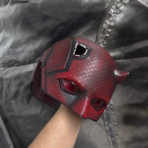 May include: A red and black Daredevil mask with a textured surface. The mask has two eye holes and a small horn on the top.