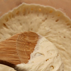 May include: A close-up of a wooden spoon stirring a light brown dough with air bubbles. The dough is in a clear glass bowl.
