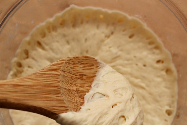 May include: A close-up of a wooden spoon stirring a light brown dough with air bubbles. The dough is in a clear glass bowl.