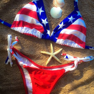May include: Red, white, and blue bikini with a star-spangled top and a solid red bottom. The bikini is lying on a brown towel with two white seashells and a starfish.
