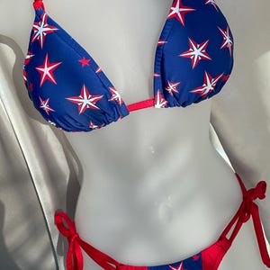 May include: A two-piece bikini set featuring a blue fabric base with a repeating pattern of red and white stars. The top has red halter straps and triangle cups, while the bottom features red side-tie closures and a matching blue star print.