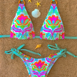 May include: A turquoise bikini with a floral print in shades of pink, orange, green, and blue. The bikini top has a halter style with gold rings and ties at the neck and back. The bikini bottom has ties on the sides.