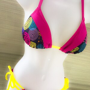 May include: A pink and black bikini with a colorful polka dot pattern. The top has adjustable straps and a tie closure in the back. The bottom has yellow ties on the sides.