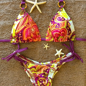 May include: A purple and white paisley patterned bikini with a triangle top and a high-cut bottom. The bikini is on a brown towel with white starfish.