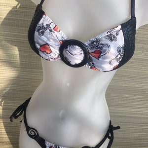 May include: Black and white bikini with a heart print and black trim. The top has a large black ring detail and the bottom has two smaller ring details.