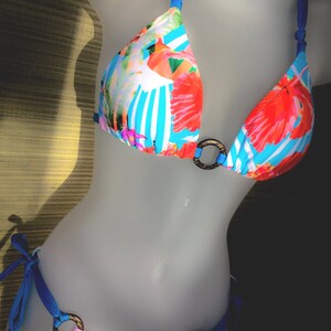 May include: A blue and white striped bikini with a floral print. The top has a halter style with a ring detail in the center. The bottom is a cheeky style with a tie side.