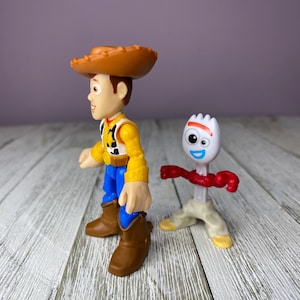 May include: Two toy figures from the animated film Toy Story. One figure is Woody, a cowboy with a brown hat, yellow shirt, blue pants, and brown boots. The other is Forky, a white spork with googly eyes, a red pipe cleaner arm, and tan legs.