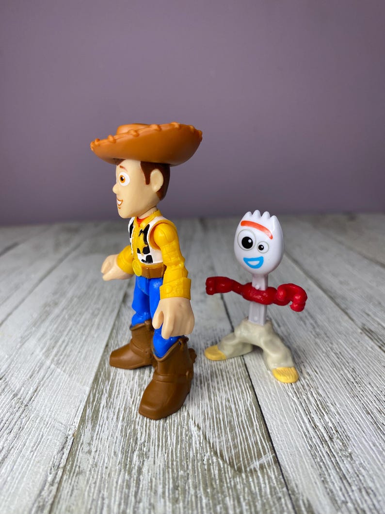 May include: Two toy figures from the animated film Toy Story. One figure is Woody, a cowboy with a brown hat, yellow shirt, blue pants, and brown boots. The other is Forky, a white spork with googly eyes, a red pipe cleaner arm, and tan legs.