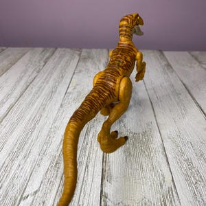 May include: A yellow and brown toy dinosaur with a long tail and detailed markings. The dinosaur is posed on a white wooden surface, with a purple background. The toy is a raptor.