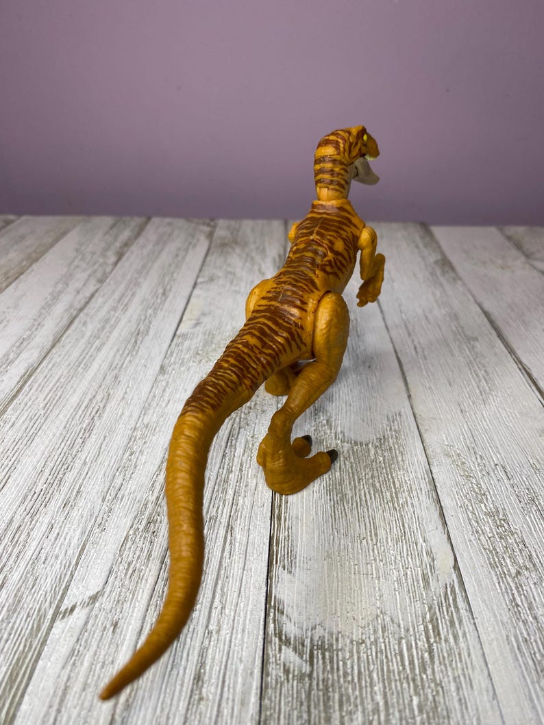 May include: A yellow and brown toy dinosaur with a long tail and detailed markings. The dinosaur is posed on a white wooden surface, with a purple background. The toy is a raptor.